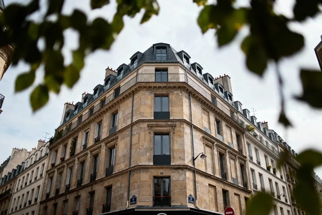 Bâtiment haussmannien en pierre, angle de rue, style parisien, feuillage flou au premier plan.