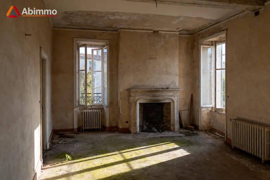 Pièce ancienne abandonnée avec cheminée, murs délabrés, fenêtres ouvertes et radiateurs sous le soleil.