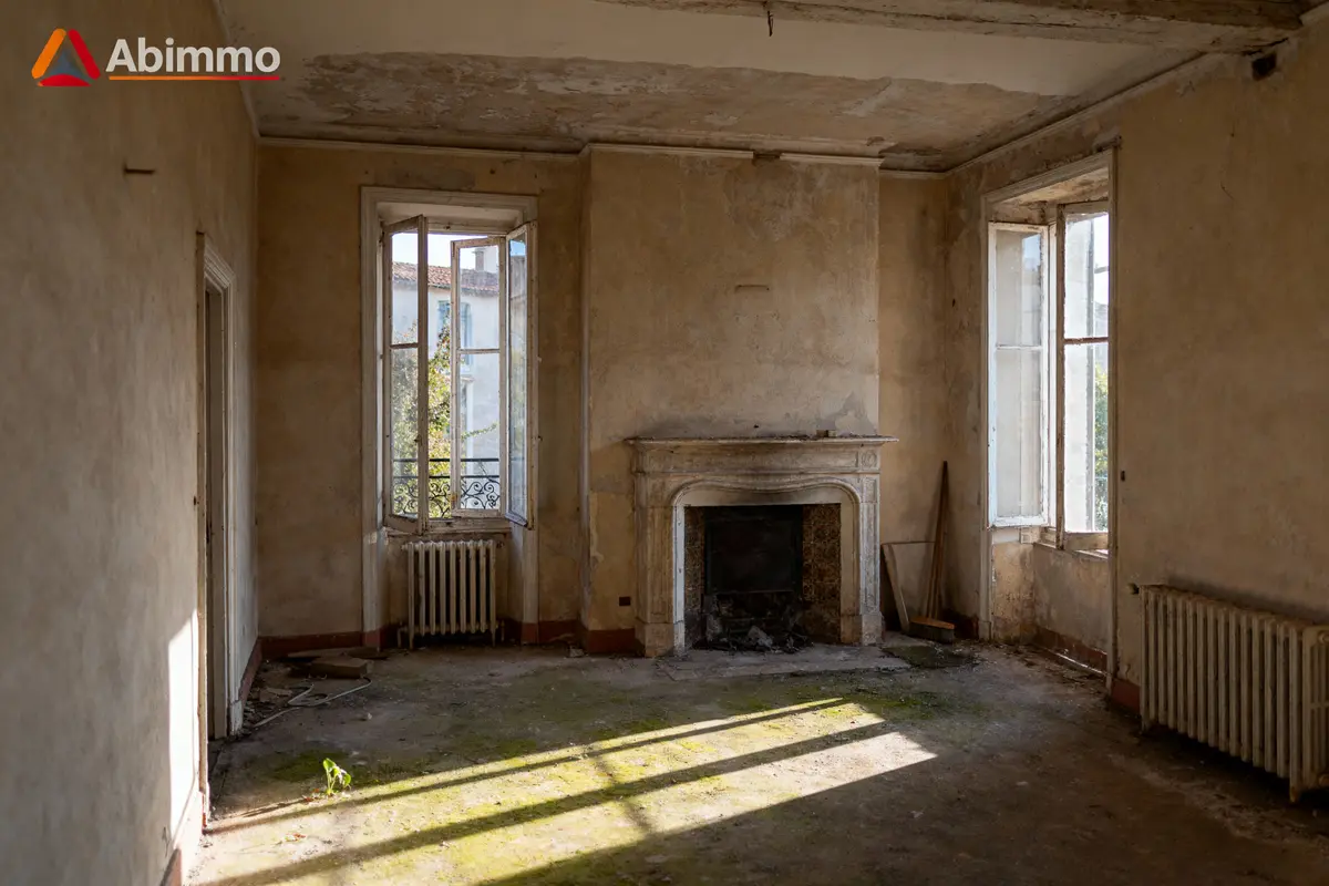 Pièce ancienne abandonnée avec cheminée, murs délabrés, fenêtres ouvertes et radiateurs sous le soleil.