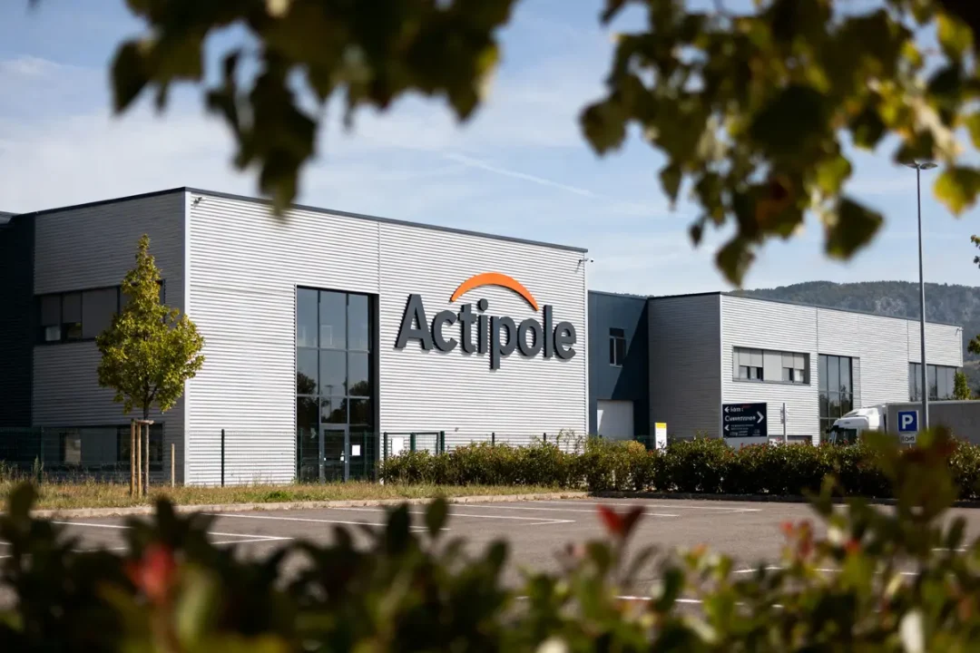 Bâtiment industriel "Actipole" avec parking et panneaux. Vue encadrée par des feuilles au premier plan.