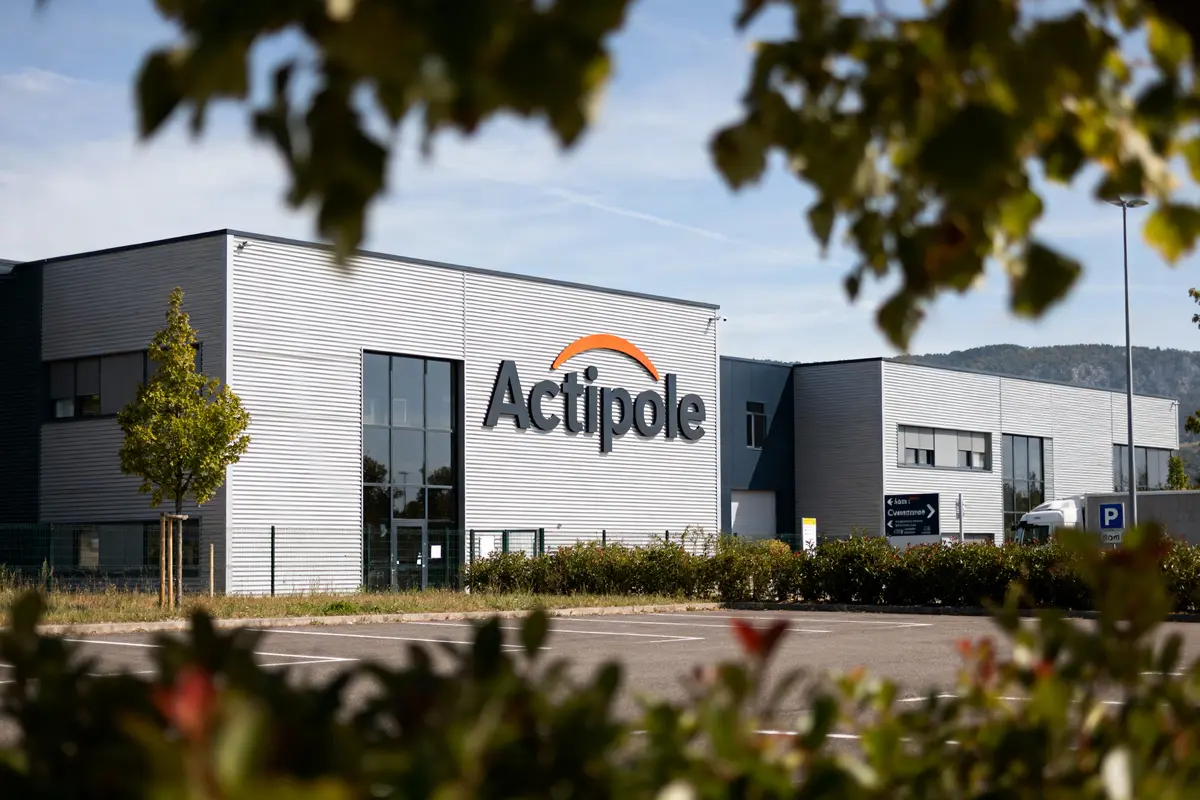Bâtiment industriel "Actipole" avec parking et panneaux. Vue encadrée par des feuilles au premier plan.