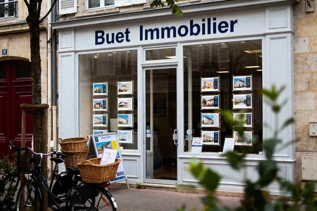Vitrine d'agence immobilière avec affiches, vélo avec panier à l'avant, rue calme.