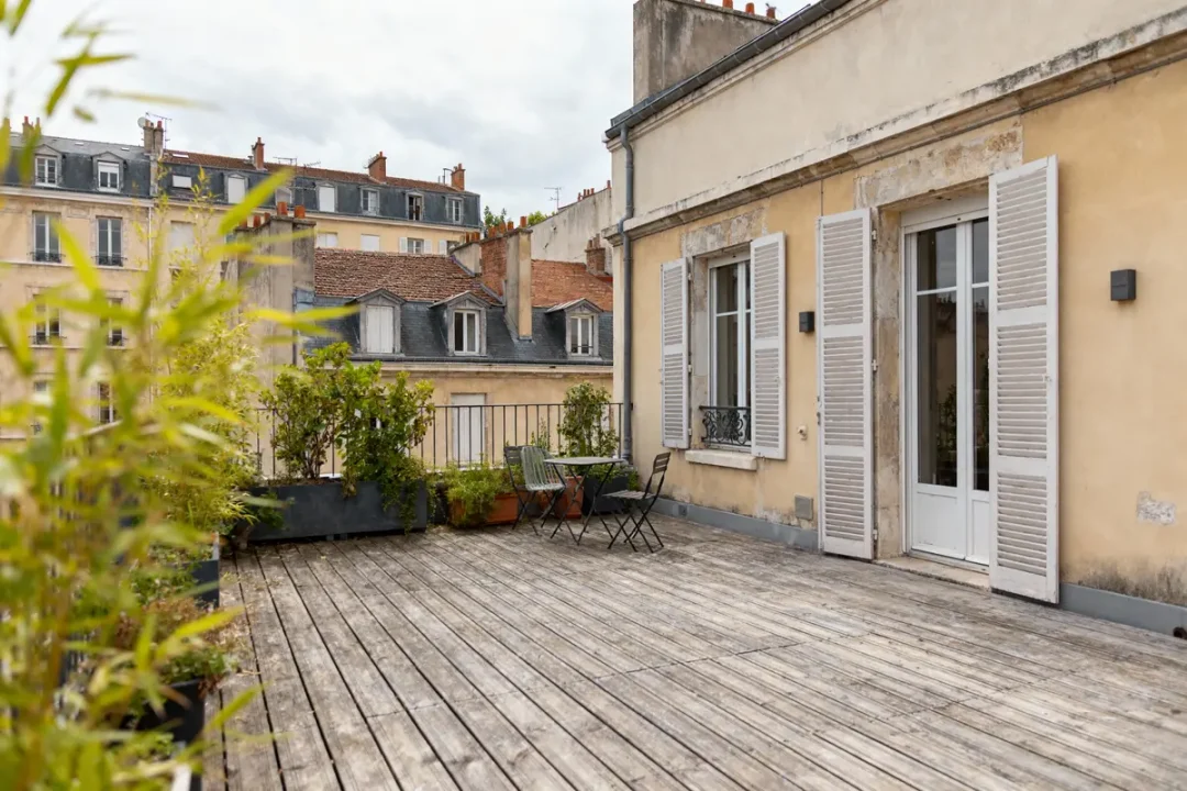Terrasse avec plantes et mobilier, surplombant toits parisiens, entourée d'anciens bâtiments résidentiels. Atmosphère paisible.