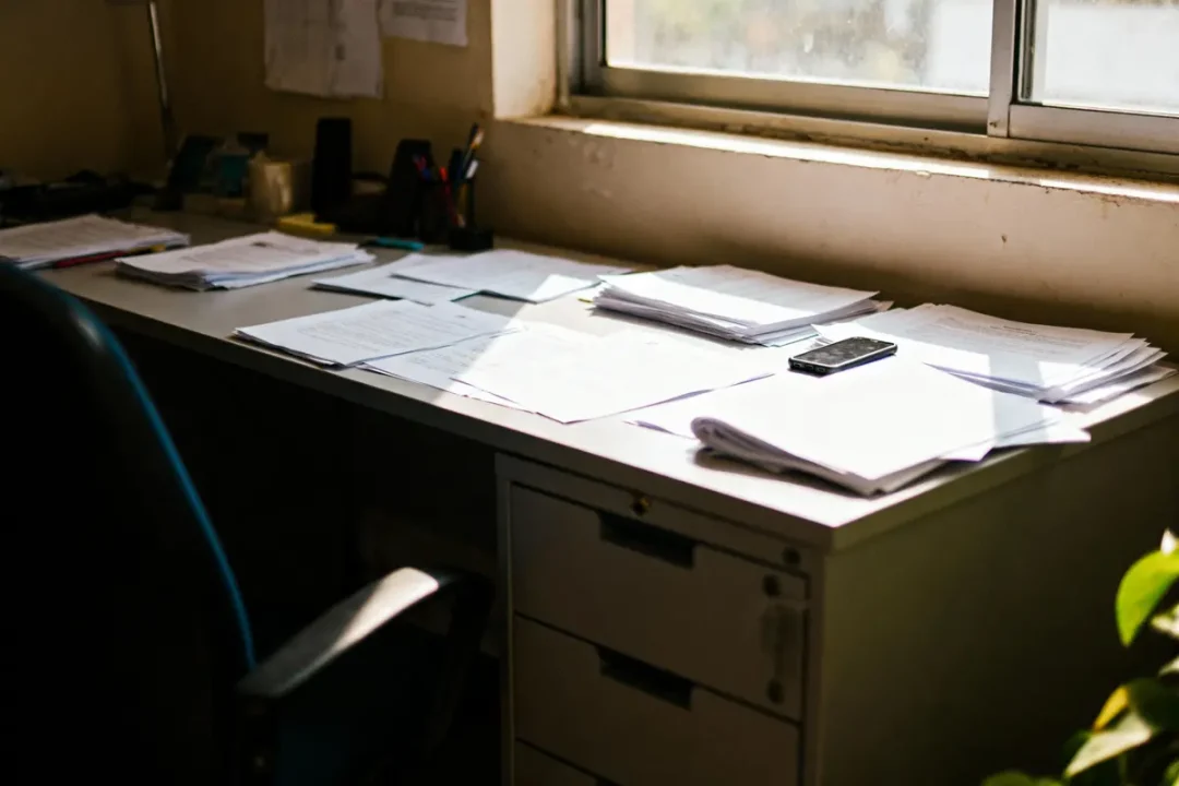 Bureau encombré de papiers sous une fenêtre, lumière naturelle, smartphone posé, quelques fournitures en arrière-plan.