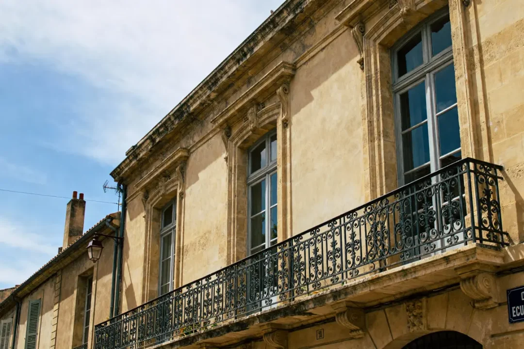 Façade de bâtiment ancien en pierre, balcons en fer forgé, fenêtres hautes, ciel bleu en arrière-plan.