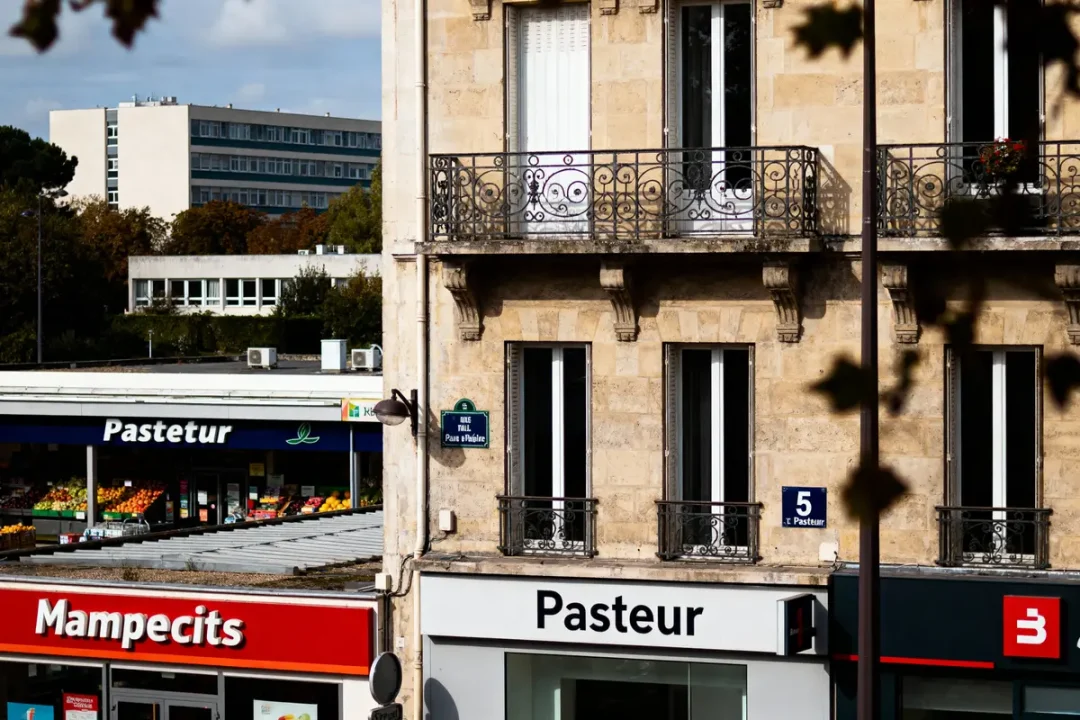Façade en pierre, immeuble ancien, balcon, devantures de magasins "Pasteur" et "Mampecits", bâtiment moderne en arrière-plan.
