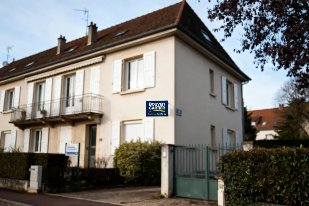 Maison avec volets blancs, balcon, jardin, panneau immobilier "Bouvencartier". Ciel bleu, arbres en arrière-plan, clôture verte.