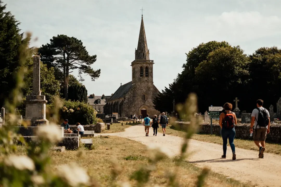 Personnes marchant sur un chemin de terre vers une église ancienne entourée de verdure et monuments.