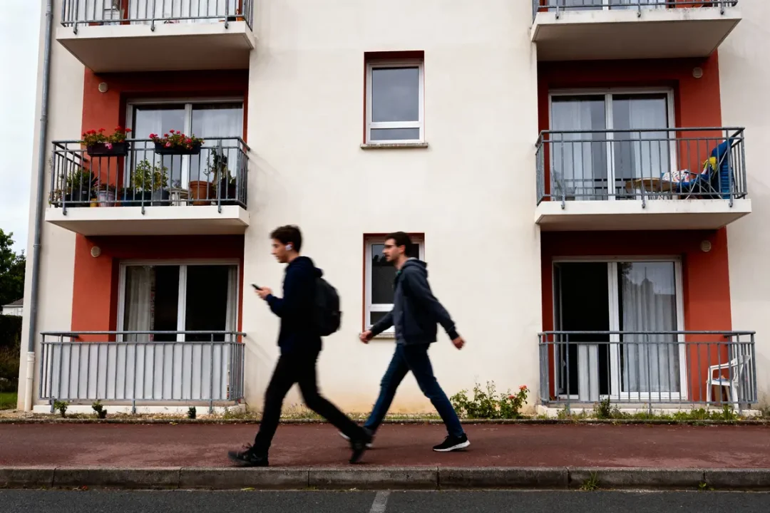 Deux personnes marchent devant un immeuble moderne avec balcons et plantes. Rue et trottoir visibles.