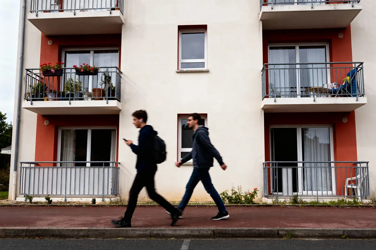 Deux personnes marchent devant un immeuble moderne avec balcons et plantes. Rue et trottoir visibles.