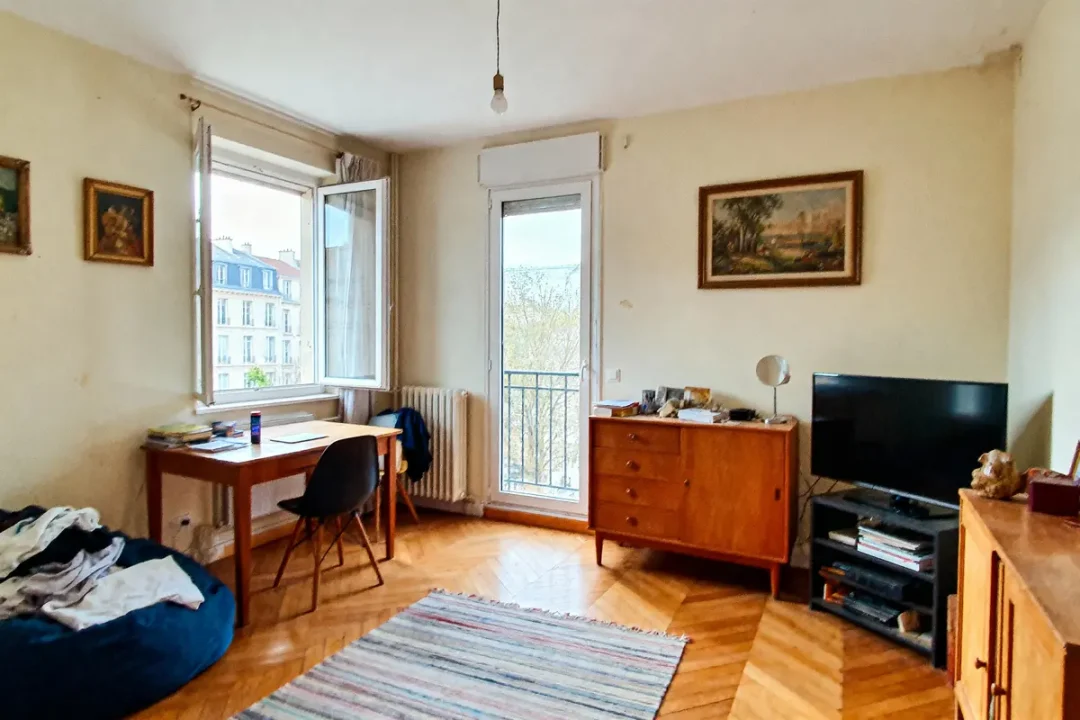 Chambre lumineuse avec bureau, chaise, commode, télévision, fenêtre ouverte, parquet en bois, tapis coloré, radiateur.