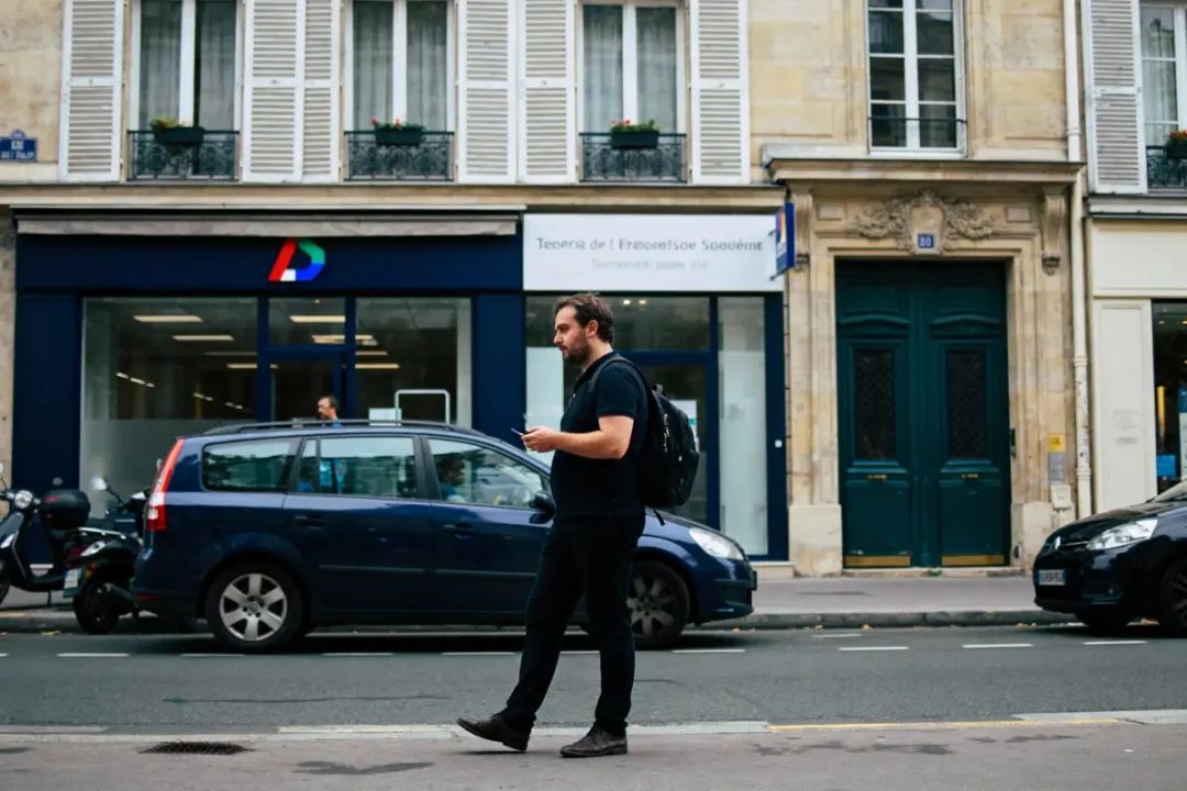 Homme marchant devant bâtiment, voiture bleue garée, rue urbaine, porte verte, volets blancs, sac à dos.