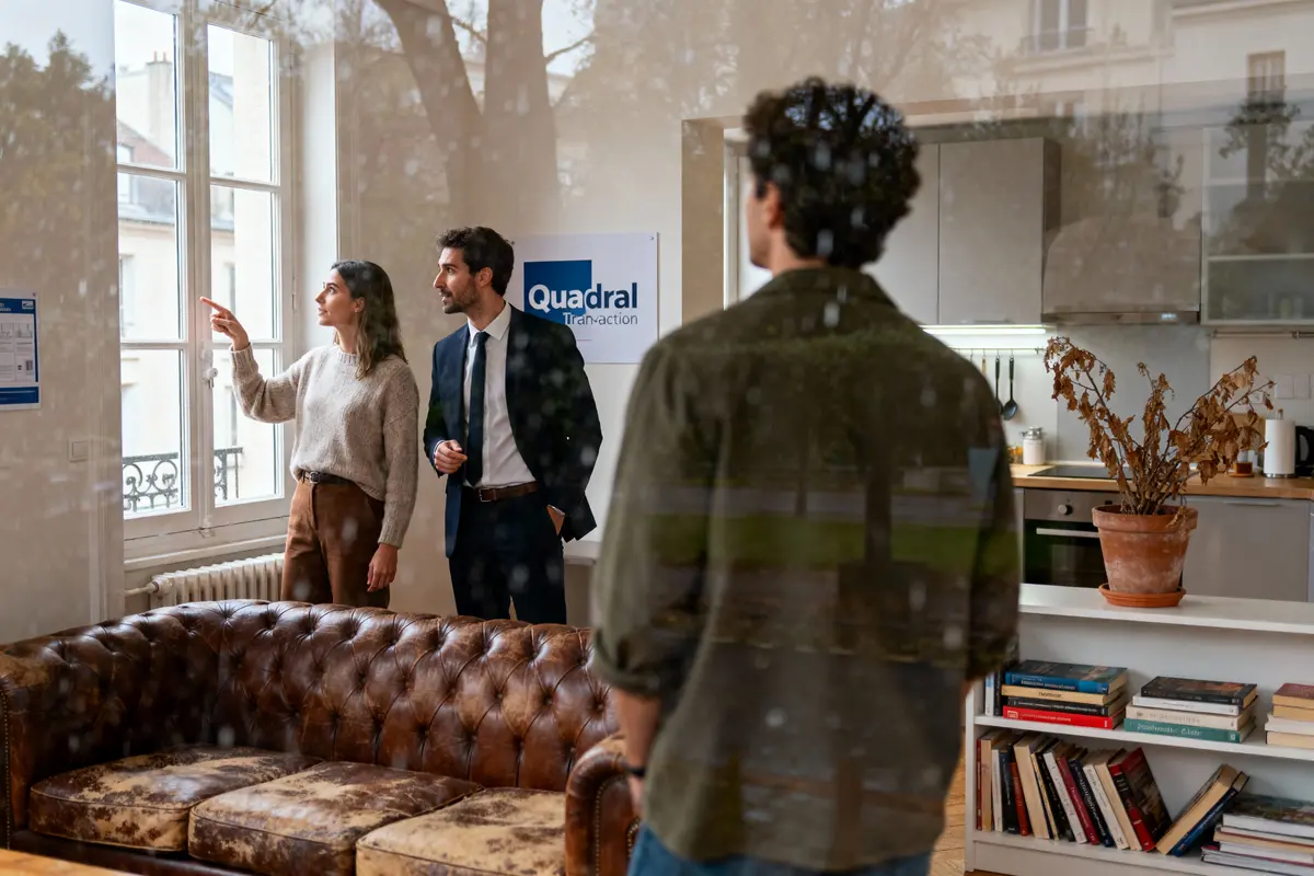 Trois personnes discutent dans un salon moderne avec canapé, bibliothèque. Reflets d'arbres sur la fenêtre.