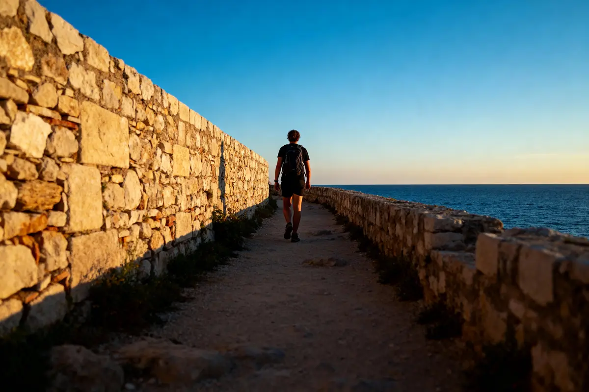 Personne marchant sur un chemin en pierre, bordant la mer au coucher du soleil.