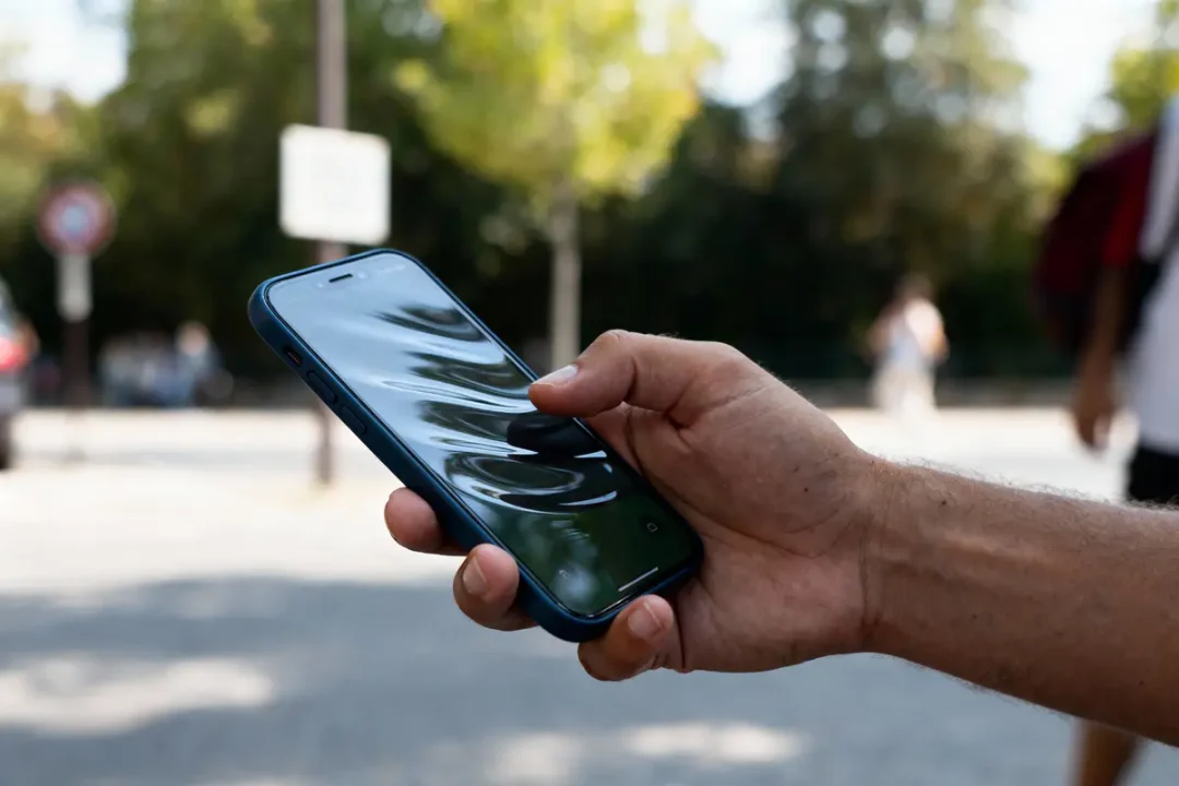 Main tenant un smartphone avec écran brillant, en extérieur, fond flou de végétation et personnes.