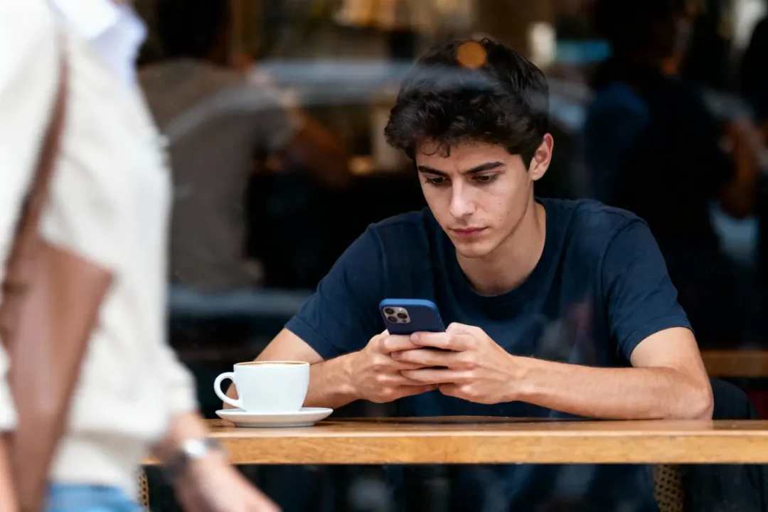 Jeune homme concentré sur téléphone portable, assis à une table avec tasse de café devant une fenêtre.