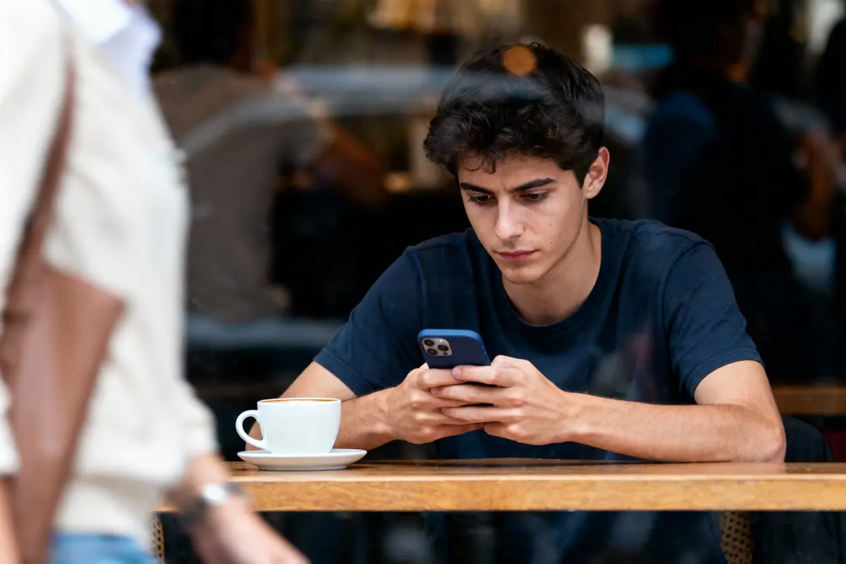Jeune homme concentré sur téléphone portable, assis à une table avec tasse de café devant une fenêtre.