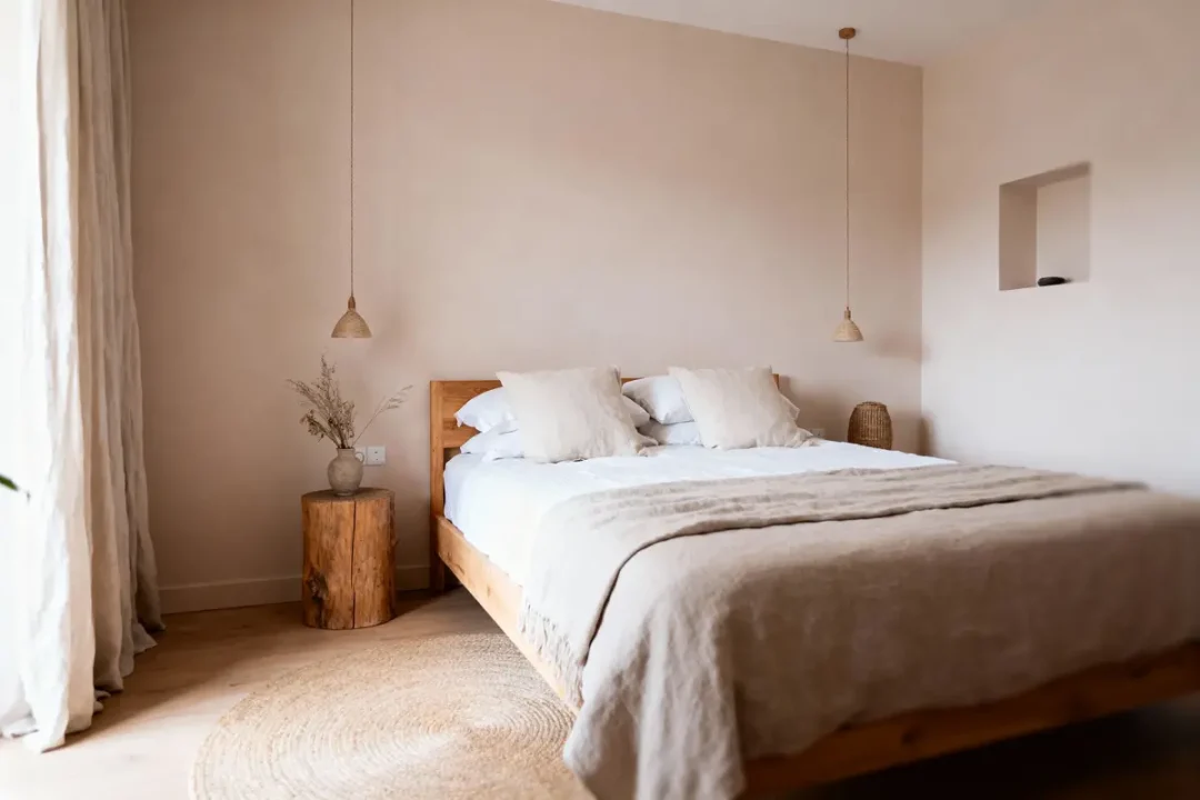 Chambre minimaliste avec lit en bois, coussins blancs, table ronde en bois, et éclairage suspendu.