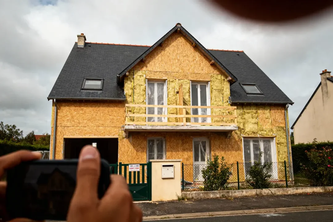 Maison en rénovation, façade avec isolation extérieure, toit en ardoise, mains tenant smartphone au premier plan.