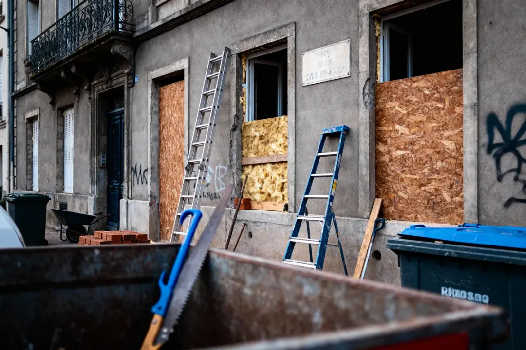 Bâtiment en rénovation, fenêtres obstruées, échelles, outils et matériaux visibles, ambiance de chantier urbain.