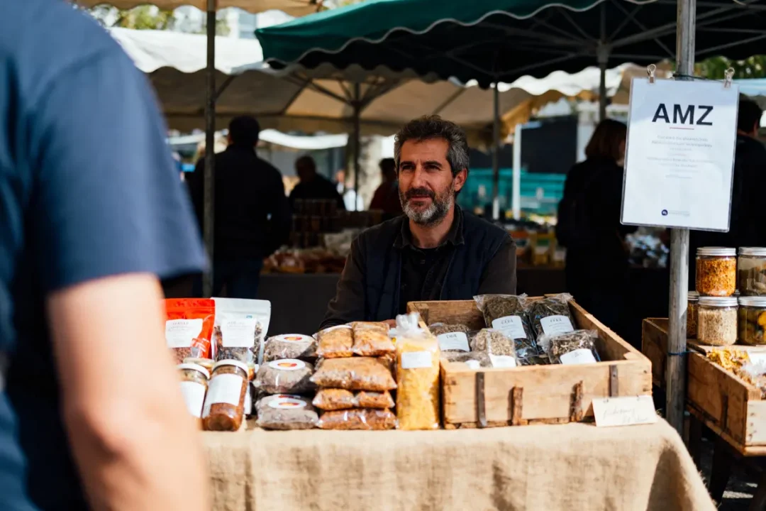 Homme vendant des produits secs sur un stand de marché en extérieur, avec clients et étals en arrière-plan.