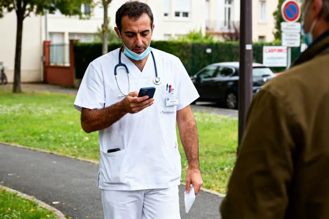 Un homme en blouse blanche consulte son téléphone en extérieur, portant un stéthoscope autour du cou.