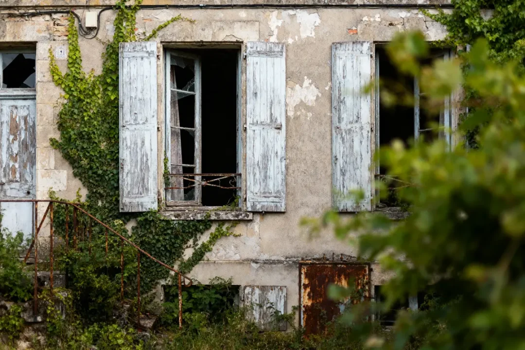 Maison abandonnée avec volets bleus vieillis, fenêtres brisées, lierre envahissant et escalier rouillé.