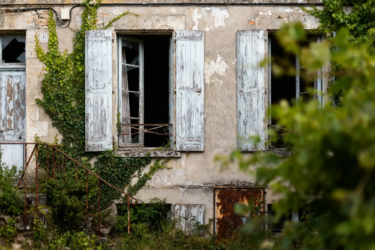 Maison abandonnée avec volets bleus vieillis, fenêtres brisées, lierre envahissant et escalier rouillé.