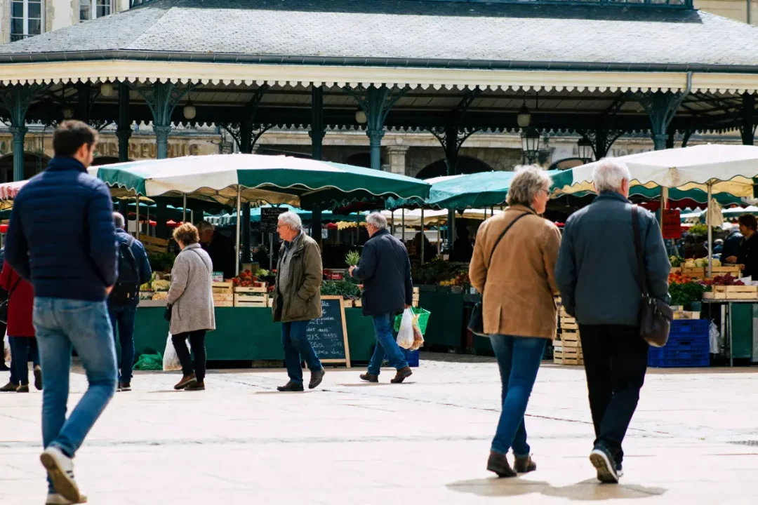 Personnes marchant dans un marché couvert, avec étals de fruits et légumes. Atmosphère animée et conviviale.