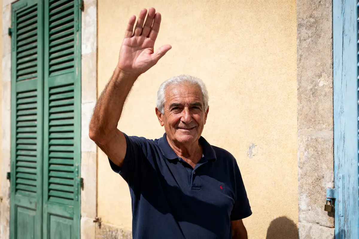 Un homme souriant, cheveux gris, lève la main devant un mur beige, près d'une fenêtre aux volets verts.