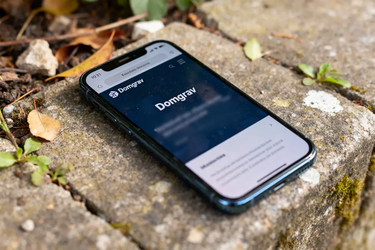 Smartphone affichant le site Domgrav sur un sol pavé avec feuilles et mousse