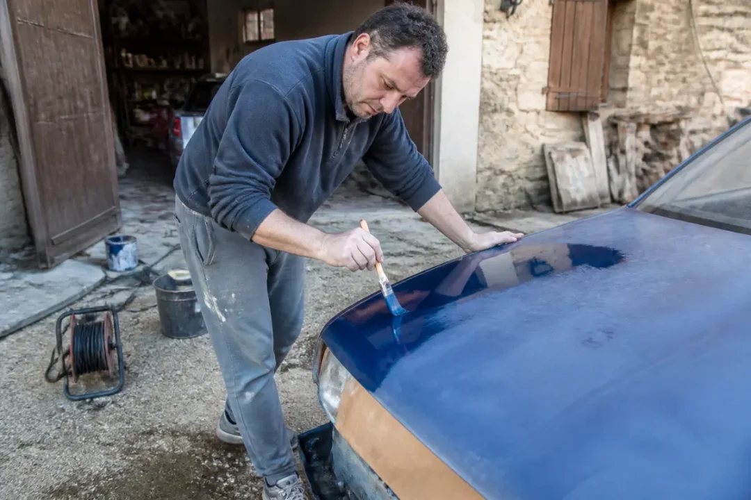 Homme repeignant une voiture bleue avec les3routes.fr peinture dans une cour ensoleillée